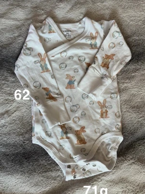 Vit omlottbody med kaniner från Lindex - Supergullig vit omlottbody från Lindex med sött kaninmönster i beige och blått. Body med långa ärmar och tryckknappar i grenen för enkel påklädning. Perfekt för små bebisar som vill vara både bekväma och stiliga.🌸 Fint använt skick. Storlek 62