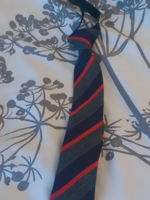 Randig slips i mörkblå och röd barn 3-5 år  - Snygg slips med diagonala ränder i mörkblått, grått och rött. Perfekt för att ge din outfit en klassisk och stilren touch. Slipsen har en smal form och passar till både skjorta och kostym.