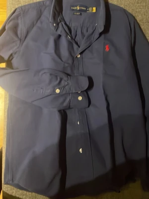 Mörkblå skjorta - Klassisk mörkblå skjorta med röd broderad logga på bröstet. Tillverkad i mjuk bomull. Storlek M, men är väldigt liten i storlek, snarare XS.