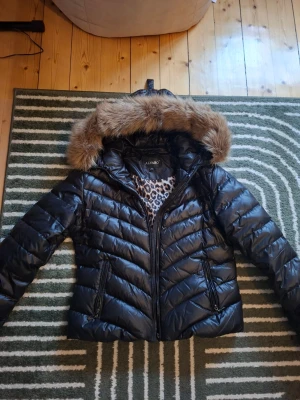Svart pufferjacka med fuskpäls - Svart pufferjacka från Alvaro med fuskpäls på huvan och leopardmönstrat foder. Skulle säga att den är i storlek 40/42.
