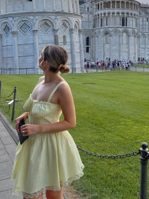 Ljusgul kort klänning med spets - Superfin ljusgul klänning med kort längd och spetsdetaljer längst ner. Klänningen har breda axelband, söt snörning vid bysten och en voluminös kjol som ger en romantisk vibe. Perfekt för varma dagar och sommarhäng.