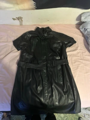 Svart skinn trenchcoat/klänning med bälte - Säljer en svart trenchcoat/klänning i skinnimitation med korta ärmar och bälte i midjan. Jackan har klassisk krage, knappar framtill och en ficka på bröstet. Perfekt för dig som vill sticka ut med en edgy look.
