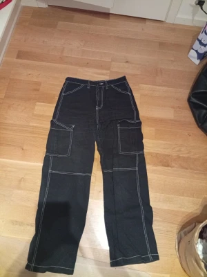 Svarta cargopants från H&M Divided - Svarta cargopants från H&M Divided i storlek 36. Byxorna har kontrasterande vita sömmar, stora fickor på sidorna och klassisk cargomodell. Passformen är loose med raka ben. Perfekt för dig som gillar streetstyle och funktionella detaljer.