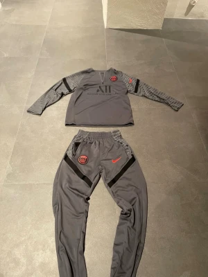 Grå PSG x Nike träningsset - Säljer ett grått PSG träningsset från Nike med långärmad tröja och matchande byxor. Setet har röda Nike-loggor och PSG-emblem, samt diskreta svarta detaljer och mönstrade partier på ärmar och ben. Materialet är mjukt och stretchigt, perfekt för fotbollsträning. Kontakta vid frågor👍👍, priset är inte hugget i sten 🪨 