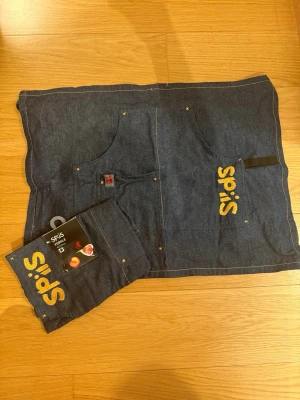 2 st Mörkblå förkläde  från Spiis - Säljer 2 st mörklblå jeans förkläde från Spiis med broderad gul text på fickan. Västen har två stora fickor framtill och kontrastsömmar i gult. 