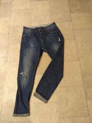 Blå jeans från Vero Moda med knappar - Snygga blå jeans från Vero Moda med knäppning framtill och slitningar på ena benet. Jeansen har klassisk femficksdesign, uppvikta benslut och kontrastsömmar. Perfekta för en avslappnad och trendig look.