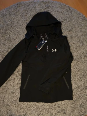 Svart vindjacka från Under Armour - Svart vindjacka från Under Armour med huva och dragkedjeförsedda fickor. Jackan har reflekterande detaljer och logga på bröstet samt meshfoder på insidan. Perfekt för blåsiga dagar och outdoor-aktiviteter.
