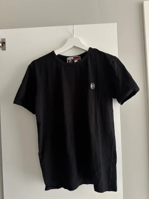 Svart t-shirt från Plein Sport - Svart t-shirt från Plein Sport med rund hals och liten logotyp på bröstet. Stilren och enkel design, perfekt för dig som gillar sportiga och cleana plagg. Materialet är mjuk bomull som känns skönt mot huden. Passformen är normal och ärmarna är korta.