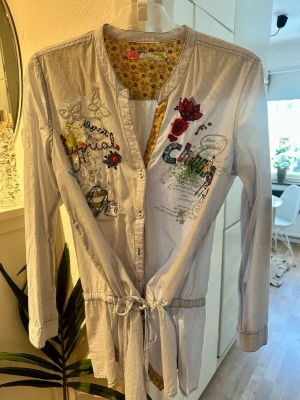 Broderad skjorta från Desigual XL - Unik långärmad skjorta från Desigual i ljusblått med tunna ränder, broderade fjärilar, blommor och färgglada detaljer. Skjortan har knappar framtill, knytband i midjan och coola tryck och broderier på bröstet. Insidan av kragen har ett blommigt mönster.