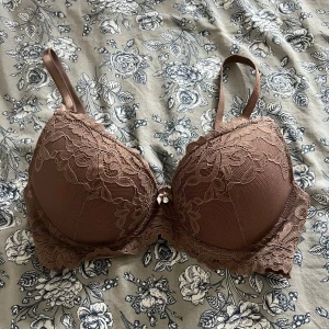 Brun spets-bh  - Oanvänd. Säljer en brun bh med vacker spets och liten rosett framtill. Bh:n har vadderade kupor och justerbara axelband. Perfekt för dig som vill ha något både snyggt och bekvämt. Ej push