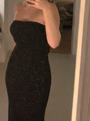 Glittrig svart klänning - Supersnygg svart bandeau-klänning i glittrigt tyg. Klänningen är figurnära och stretchig så den sitter väldigt fint på kroppen. Den slutar några centimeter över mina anklar och har en liten slit på sidorna. För frågor eller mer bilder är det bara att höra av sig i DM🖤