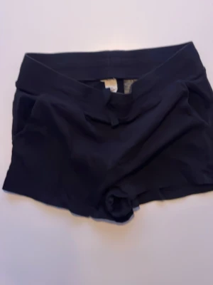 Svarta midwaist träningsshorts - Säljer ett par svarta midwaist shorts som är perfekta för träning. De har en enkel och stilren design med korta ben och mjukt syntetmaterial som sitter skönt mot huden. Passar dig som vill ha bekväma och funktionella shorts till gymmet eller löprundan.