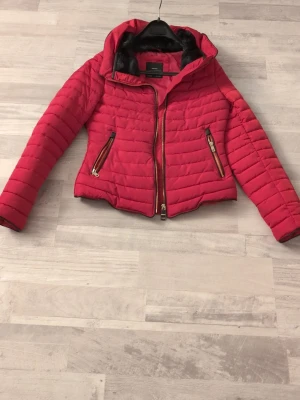 Röd pufferjacka från Zara Basic - Säljer en snygg röd pufferjacka från Zara Basic med svarta fuskpälsdetaljer i kragen. Jackan har guldiga dragkedjor på fickorna och framtill, quiltad design och normal passform. Perfekt för kyliga dagar när du vill sticka ut lite extra.