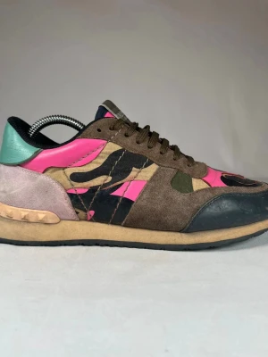 Valentino Rockrunner sneakers med camomönster - Säljer ett par Valentino Rockrunner sneakers med camouflagemönster i brunt, grönt, svart och neonrosa. Skorna har detaljer i mocka, skinn och textil samt snörning och rund tå. Sulan är beige med nitar på hälen och inslag av mintgrönt och ljusrosa baktill.