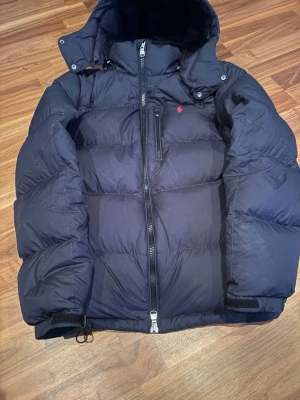 Marinblå pufferjacka från Polo Ralph Lauren - Säljer en marinblå pufferjacka från Polo Ralph Lauren med huva och dragkedja. Jackan har två fickor med dragkedja och den klassiska röda Polo-loggan på bröstet. Perfekt för kalla dagar och riktigt snygg till streetwear-stilen. Den har ett litet hål i ryggen men inget som står ut så mycket 