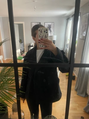 Svart/mörkblå  kappa från design by si - Säljer en svart/mörkblå  kappa från design by si och bred krage. Jackan har stora fickor framtill och dekorativa knappar vid ärmsluten. Klassisk och tidlös modell i ullblandning som passar perfekt till höst och vinter. Produkten är använd fåtal gånger men inga defekter