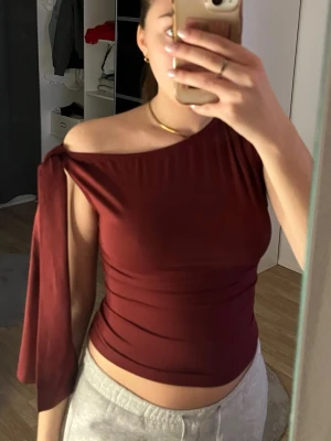 Vinröd oneshoulder topp med knyt - Säljer en vinröd oneshoulder topp med snygg knyt-detalj på ena axeln. Toppen är tight och har en croppad passform.