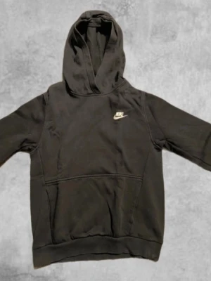 Svart Nike hoodie - Svart hoodie från Nike med klassisk vit logga på bröstet. Tröjan har huva, lång ärm och en stor magficka framtill. Tillverkad i mjuk bomullsmix, perfekt för chill eller streetwear. Enkel och stilren design som funkar till det mesta. Storlek 147-158.