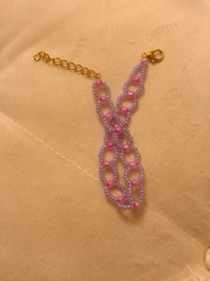 Lila och rosa pärlarmband med guldlås - Handgjort armband med små lila och rosa pärlor i ett snyggt, öppet mönster. Armbandet har ett guldfärgat kedjelås som gör det enkelt att justera storleken. Perfekt accessoar för att ge din outfit lite extra färg och personlighet.