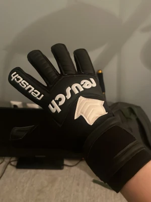 Svarta målvaktshandskar från Reusch - Säljer ett par svarta målvaktshandskar från Reusch med vit logga och detaljer. Handskarna har greppig insida och bred kardborrestängning vid handleden. Perfekta för dig som vill ta ditt spel till nästa nivå på fotbollsplanen. Helt oanvända och plasten sitter fortfarande kvar på latexen. Storlek 10. 