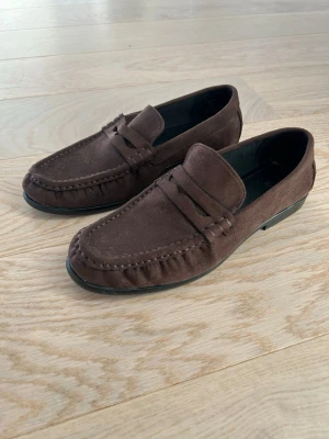 Bruna loafers i mocka  - Snygga loafers i mörkbrun mockaimitation från H&M i strl 38. Använda en gång och är i mycket bra skick.