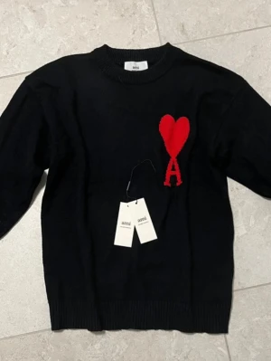 Svart  tröja från Ami Paris - Svart stickad tröja från Ami Paris med röd hjärtlogga och bokstaven A på bröstet. Tröjan har rund hals, ribbade muddar och lång ärm. Perfekt för dig som vill ha en stilren men ändå unik look.