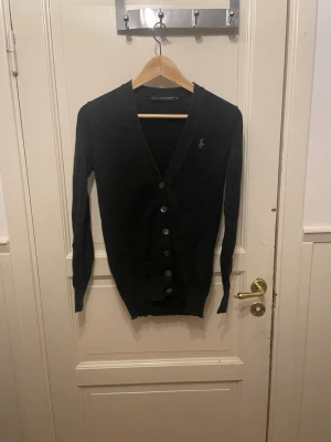Svart kofta från Ralph Lauren - Svart kofta från Ralph Lauren med lila broderad logga på bröstet. Koftan har v-ringning, långa ärmar och knappar framtill. Materialet är mjukt och stickat, perfekt för lager-på-lager under höst och vinter. Snygg och klassisk design som funkar till många stilar.