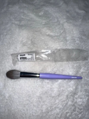 Lila sminkborste från DAAR - Snygg sminkborste från DAAR med lila handtag och mjuka, täta borststrån. Borsten har en metallisk silverfärgad övergång mellan borst och handtag. Perfekt för applicering av puder, bronzer eller rouge. Modellnummer 03 står på handtaget.