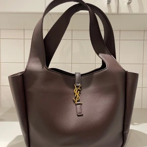 Brun handväska från Saint Laurent - Snygg brun handväska från Saint Laurent i slät skinn med guldigt spänne framtill. Väskan har breda axelremmar och rymligt innerfack med dragkedja. Perfekt för dig som vill ha en stilren och lyxig accessoar med ikonisk design.