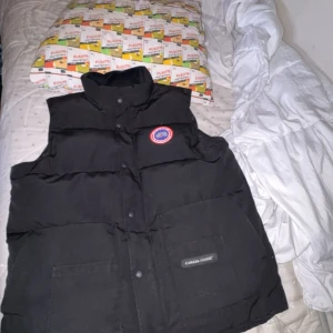 Svart dunväst från Canada Goose - Svart dunväst från Canada Goose med klassisk logga på bröstet och hög krage. Västen har två stora fickor framtill, stängs med både dragkedja och knappar. Perfekt för lager-på-lager och riktigt varm med sin quiltade design.