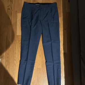 Blå kostymbyxor Tiger of Sweden 52( M/L) - Snygga blå kostymbyxor från Tiger of Sweden i storlek 52. Byxorna har klassisk rak passform, pressveck och bälteshällor. Perfekta till en dressad look och tillverkade i ett elegant tyg med diskret struktur.