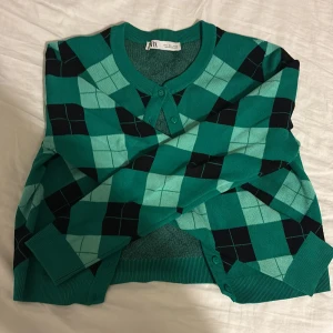 Grön rutig kofta från Zara - Snygg grön kofta från Zara med rutigt mönster i svart och ljusgrönt. Koftan har rund halsringning, knappar framtill och ribbade muddar. Perfekt för dig som vill sticka ut med en färgglad och trendig look.