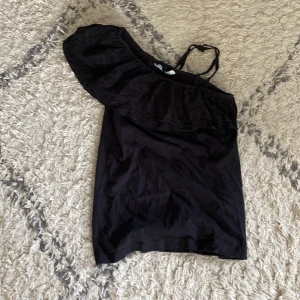 Svart offshoulder topp med volang - Snygg svart topp med offshoulder-design och bred volang över ena axeln. Toppen har smala axelband och är tillverkad i mjukt bomullstyg. Perfekt för dig som vill ha en trendig och stilren look.