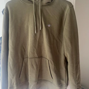 Olivgrön hoodie från Gant i bomull - Olivgrön hoodie från Gant med broderad logga på bröstet. Klassisk känguruficka framtill, justerbar huva med snören och ribbade muddar. Skön och stilren modell i mjuk bomull, perfekt för chill dagar.