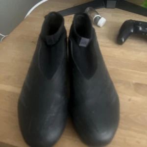 Svarta Adidas Predator fotbollsskor - Säljer ett par svarta Adidas Predator fotbollsskor med klassisk design och diskreta ränder på sidan. Skorna har rund tå, platt sula med fasta dobbar och är tillverkade i syntetmaterial. Perfekta för dig som vill dominera på planen.