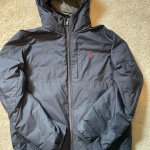 Mörkblå pufferjacka från Polo Ralph Lauren - Snygg mörkblå pufferjacka från Polo Ralph Lauren med huva och röd broderad logga på bröstet. Jackan har en rejäl dragkedja med Polo-detalj, quiltad insida och praktiska fickor. Perfekt för kalla dagar och stilren look.