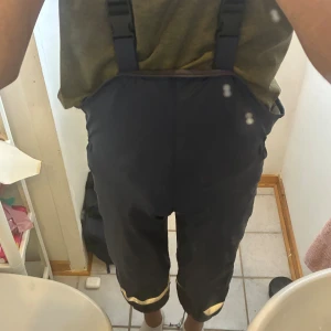 Svarta täckbyxor med hängslen - Svarta täckbyxor med justerbara hängslen och reflexdetaljer vid bensluten. Byxorna är trekvartsmodell och perfekta för utomhusaktiviteter under kallare dagar. Praktisk och bekväm passform. Hör av dig för mer information och bilder 