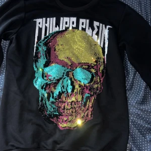 Svart Philipp Plein sweatshirt med paljetter - Cool svart sweatshirt från Philipp Plein med stort glittrigt dödskalletryck i guld, turkos och rosa paljetter framtill. Tröjan har rund hals, långa ärmar och ribbade muddar. Märkets logga i metall på ena ärmslutet. Perfekt statement piece för dig som gillar att sticka ut.