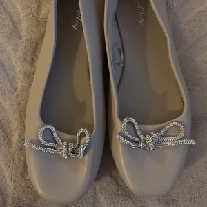 Beige ballerinaskor med strassrosett - Snygga beige ballerinaskor från Alley med fyrkantig tå och dekorativa rosetter i glittrande strass framtill. Skorna har platt sula och är tillverkade i syntetmaterial, perfekta för en chic och trendig look.