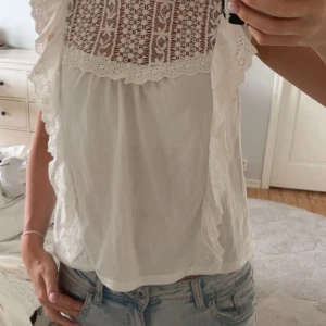 Vit blus med spets och volang - Supersöt vit blus med spets upptill och volangdetaljer längs axlarna. Blusen är ärmlös och har ett romantiskt mönster i spetsen samt broderade kanter. Perfekt för dig som gillar en feminin och fräsch stil. Passar till jeans eller kjol.
