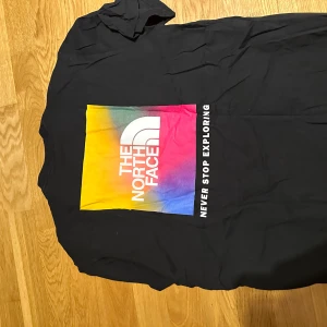Svart The North Face t-shirt med färggrafik - Svart t-shirt från The North Face i bomull med stor färgglad grafisk print på ryggen i gult, rosa och blått samt vit logga och texten 'Never Stop Exploring'. Klassisk passform och korta ärmar. Liten vit logga framtill på bröstet.