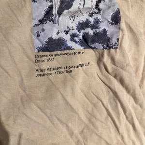 Beige t-shirt med Hokusai-print - Beige t-shirt med två konstmotiv av Katsushika Hokusai tryckta framtill. Ena trycket visar tranor på snötäckt tall, det andra en våg. T-shirten är kortärmad och har en avslappnad passform. Materialet är mjuk bomull, perfekt för en chill och konstnärlig vibe.