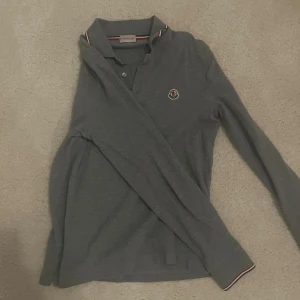 Grå långärmad pikétröja Moncler - Säljer en stilren grå långärmad pikétröja från Moncler med klassisk krage och knäppning framtill. Tröjan har Moncler-logga på bröstet och diskreta ränder i rött och vitt på kragen och ärmsluten. Perfekt för dig som gillar en clean och exklusiv look. Köpt på vestiarie collective.