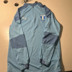 Malmö FF långärmad träningströja Puma - Ljusblå långärmad fotbollströja från Puma med Malmö FF-logga på bröstet och vita Puma-detaljer. Tröjan har dragkedja vid halsen och mörkare blå partier på ärmarna. Tillverkad i lätt och funktionellt material, perfekt för träning eller match.