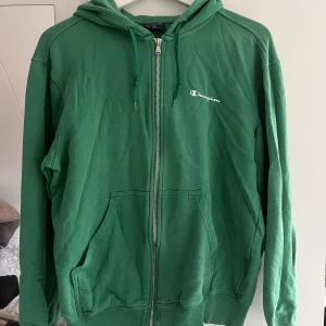 Grön hoodie med dragkedja från Champion - Grön hoodie från Champion i storlek L med dragkedja framtill och huva med snörning. Tröjan har två stora fickor på magen och broderad Champion-logga på bröstet. Perfekt för chill dagar eller när du vill ha en sportig look. Passar både herr (M) och dam (M-L)