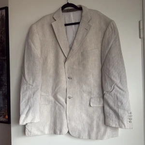 Randig ljusbeige kavaj från Collezione - Snygg ljusbeige kavaj från Collezione i italiensk linne. Kavajen har diskreta vita ränder, klassisk krage, två knappar framtill och fyra knappar vid ärmslutet. Fodret är i satin och insidan har flera fickor. Perfekt för dig som gillar stilrena detaljer.