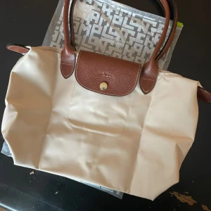 Beige handväska från Longchamp - Snygg beige handväska från Longchamp med bruna skinnhandtag och lock. Väskan har guldfärgad knapp och dragkedja upptill. Perfekt storlek för det viktigaste och klassisk design som funkar till många stilar.