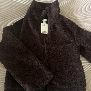 Brun teddyjacka från H&M XXS - Mysig brun teddyjacka från H&M i storlek XXS. Jackan har dragkedja framtill, hög krage och två stora fickor. Perfekt för kyliga dagar när du vill ha en chill och trendig look.