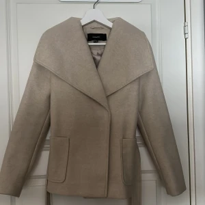 Beige kappa från Vero Moda XS - Stilren beige kappa från Vero Moda i storlek XS. Jackan har bred krage, stora fickor framtill och bälte i midjan för snygg passform. Perfekt för dig som gillar en klassisk look med modern twist.