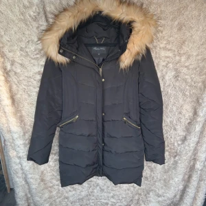 Svart quiltad vinterjacka med fuskpäls - Svart vinterjacka från Happy Holly med fluffig beige fuskpäls på huvan. Jackan har guldiga dragkedjor på fickorna och framtill. Varm och skön
